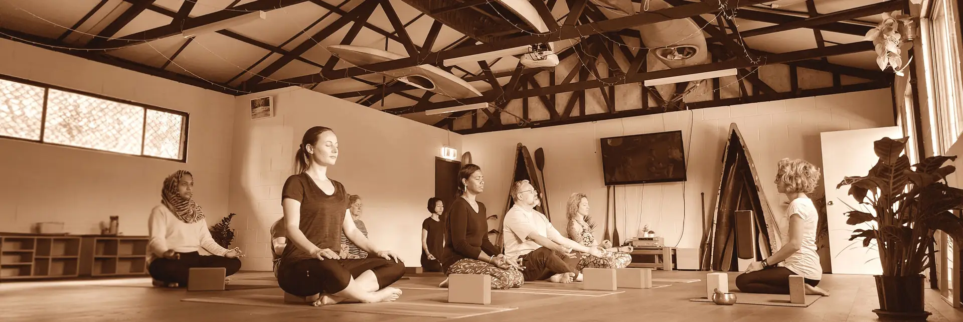 Yoga class in session at River Canoe Club Marrickville. Participants are seated in a peaceful studio space by the Cooks River, with teacher Stanja guiding in the background.